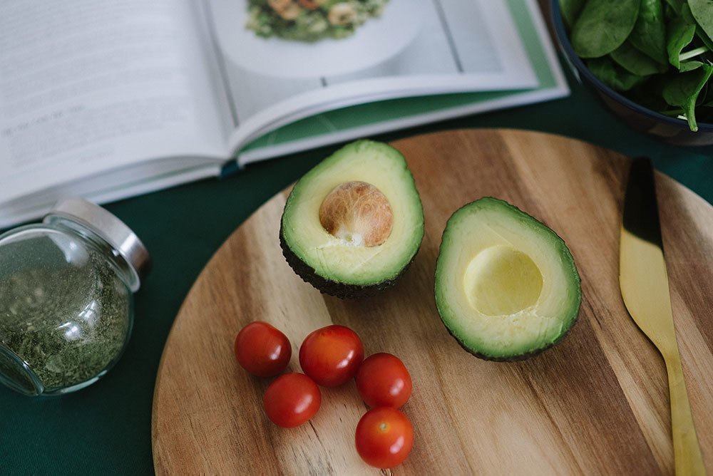 One avocado and cherry tomatoes on a wooden chopping board, next to a recipe book.